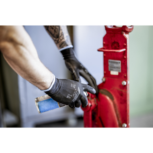 Eine Person mit NITRAS Nylonhandschuhen, schwarz, PU-beschichtet von AS Arbeitsschutz GmbH reinigt oder schmiert mit einer blauen Sprühdose ein rotes Maschinenteil aus Metall. Zu sehen sind lediglich Arme und Hände.