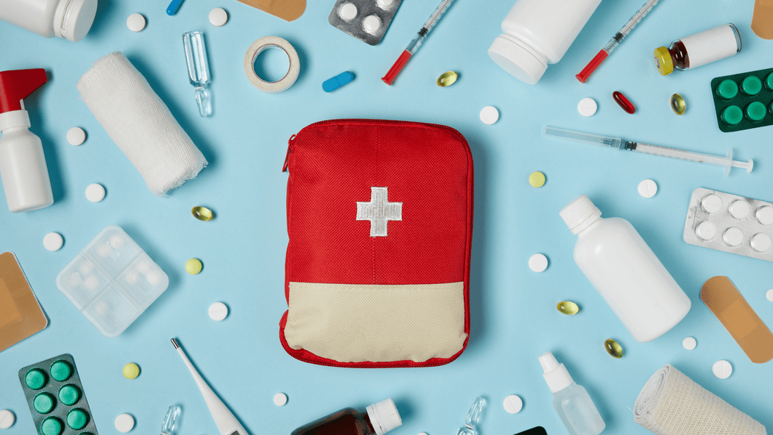 Rote Erste-Hilfe-Tasche mit weißem Kreuz, umgeben von Medikamenten, Spritzen und Verbandsmaterial auf blauem Hintergrund