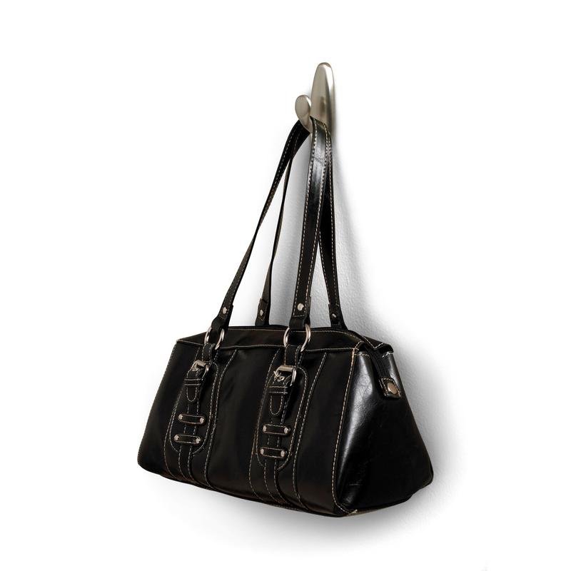 À un crochet en métal Command™ "classique" de 3M Deutschland GmbH est suspendu un sac à main en cuir noir avec des poignées doubles et des boucles décoratives. Le sac a des coutures blanches et une surface légèrement brillante.