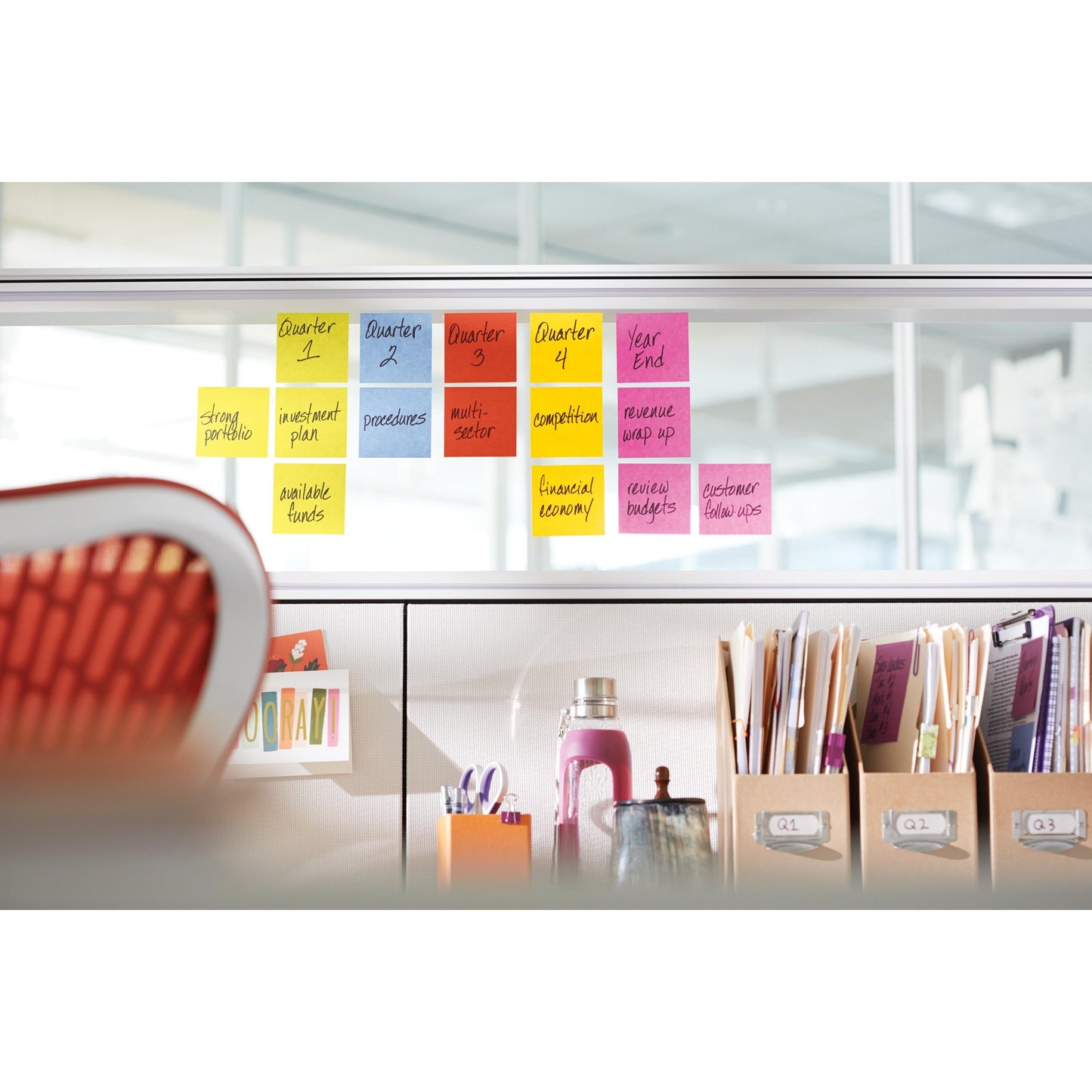 Une série de Post-it® Super Sticky Notes de 3M Deutschland GmbH, 76 mm x 76 mm, 90 feuilles/bloc, 100% PEFC, est disposée sur une cloison en verre dans le bureau au-dessus de fournitures organisées, de classeurs et de livres avec une chaise orange devant.