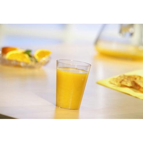 Un gobelet Starpak 50 rempli de jus d'orange (PS transparent) est posé sur une table claire, avec une assiette floue de tranches d'orange et une serviette jaune avec du pain ou des crackers en arrière-plan.