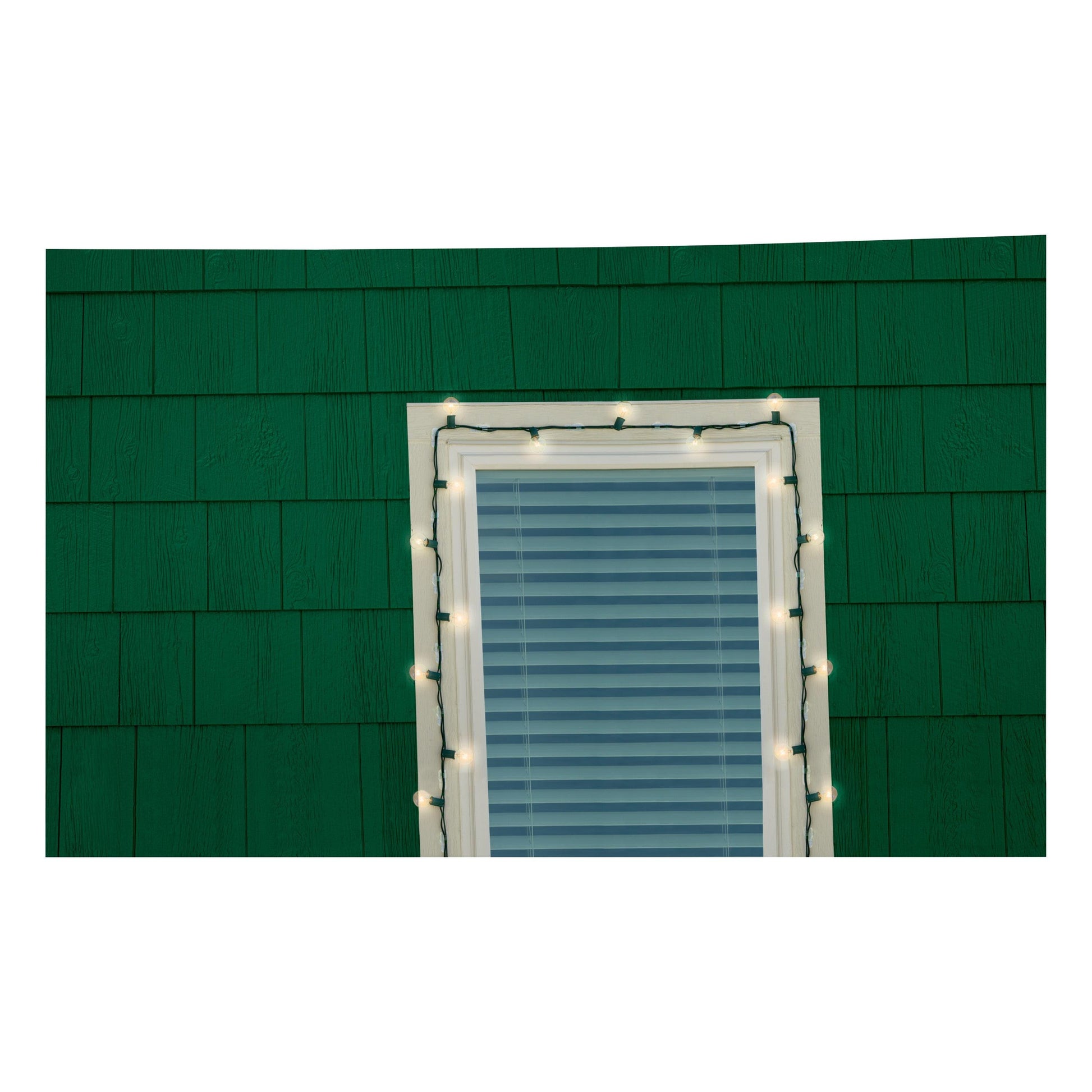 Eine grüne Wand zeigt ein Fenster mit geschlossenen Jalousien, eingerahmt von weißen Lichterketten, die mit 3M Deutschland GmbH Command™ Outdoor Lichter-Clips, 17017CLR-AWPU (8 Clips + 10 Streifen) aufgehängt wurden. Die Lichter sind eingeschaltet.