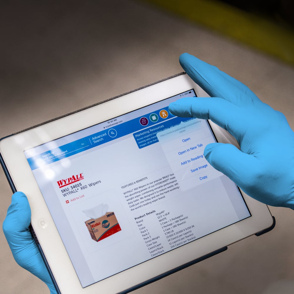 Eine Person, die KleenGuard® G10 FleX Nitril-Handschuhe von Kimberly-Clark GmbH trägt, benutzt ein Tablet, auf dem Produktdetails und die Verpackung dieser 24 cm langen, beidhändigen Nitril-Handschuhe auf einer Webseite angezeigt werden.