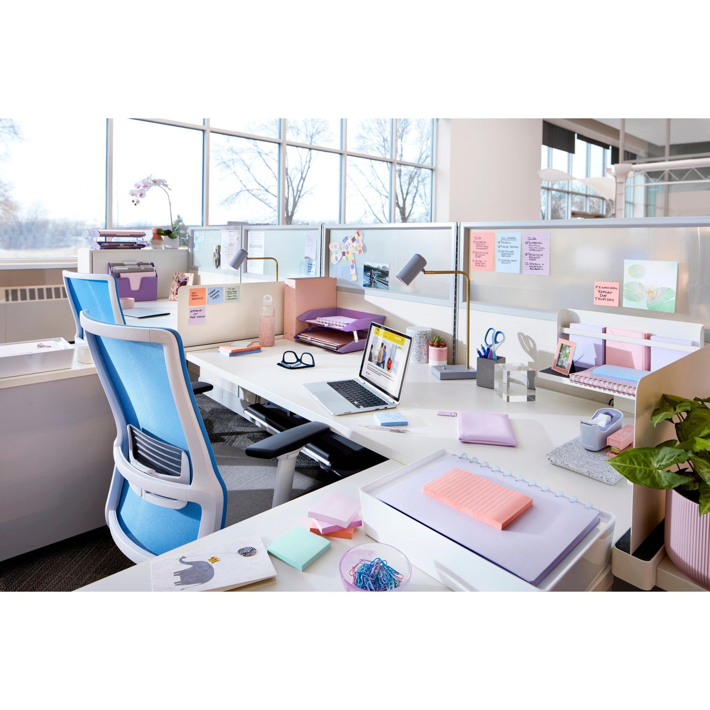 Un bureau moderne avec des bureaux blancs, des chaises ergonomiques bleues, des ordinateurs portables, des fournitures, des effets personnels, des plantes en pot et des Post-it® Super Sticky Notes (76x76 mm, 90 feuilles/bloc, 100% PEFC) de 3M Deutschland GmbH sur les cloisons; à l'extérieur, un paysage enneigé.