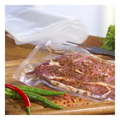 Un steak cru mariné est mis sous vide dans un sac transparent Starpak 100 à bord scellé (PA/PE) avec une machine à vide à cloche sur une planche en bois, avec des piments, des grains de poivre rose, des légumes-feuilles et une fourchette en arrière-plan.