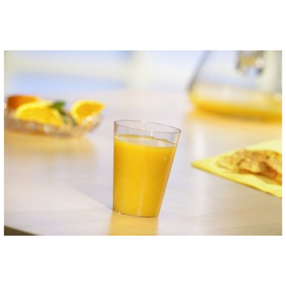 Un gobelet Starpak 50, PS transparent de Starpak, rempli de jus d'orange, est posé sur une table claire. Derrière se trouvent des tranches d'orange sur une assiette et un objet jaune flou, peut-être de la nourriture, sur une serviette jaune.