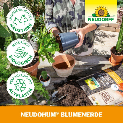 Une personne rempote une plante sur une table avec de la terre, des plantes de jardin en pot, des outils de jardin, des sacs de terreau et un sac de NeudoHum BlumenErde du Neudorff Shop. À côté du logo Neudorff apparaissent les symboles d'écologie et de convivialité pour les animaux de compagnie.