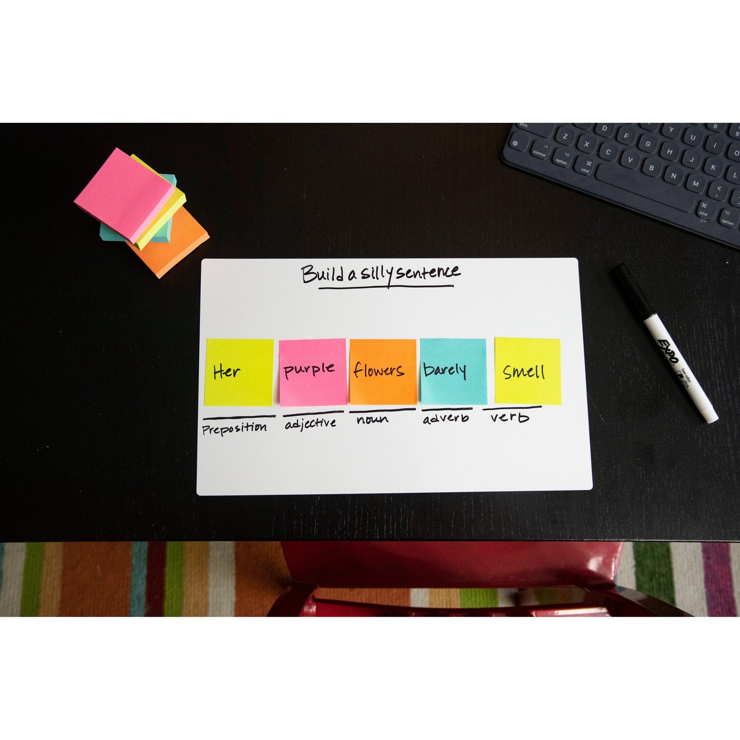 Auf einem Schreibtisch stehen Post-it® Super Sticky Z-Notes von 3M Deutschland GmbH mit den Wörtern für einen albernen Satz neben einem Whiteboard mit der Aufschrift "Build a silly sentence". Auf dem Tisch befinden sich auch ein trockener Löschstift und eine Tastatur.