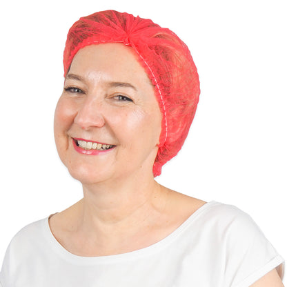 Une femme souriante dans une chemise blanche et une ARNOMED COVER PP-KLIPPHAUBE rouge vif, une charlotte jetable en non-tissé PP (52 cm, ARNOWA GmbH) se tient devant un fond blanc simple.