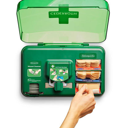 Un article vert de Orkla Wound Care AB monté au mur : distributeur de soins des plaies Cederroth (20,3x30,6x15,5 cm) avec couvercle ouvert montre des nettoyants pour plaies et des matériaux de bandage, tandis qu'une main tire un bandage du set de sachets en plastique du distributeur de pansements.