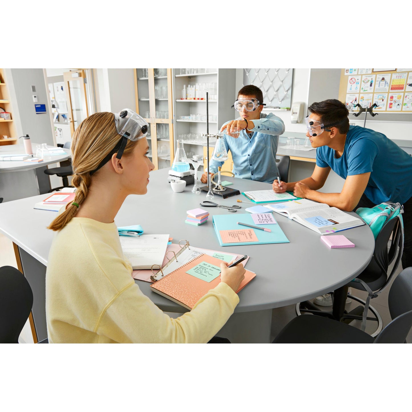 Trois étudiants avec des lunettes de protection sont assis à une table de laboratoire ronde avec des carnets, des manuels et des Post-it® Super Sticky Notes (76 mm x 76 mm, 90 feuilles/bloc, 100% PEFC) de 3M Deutschland GmbH et réalisent une expérience avec un statif et un bécher.