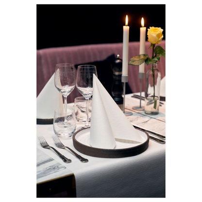 Une table à manger est élégamment dressée avec deux serviettes Duni Dunilin® 48 x 48 cm blanches, pliées en triangles sur les assiettes. Deux grandes bougies allumées et une rose jaune dans un vase en verre décorent l'arrière-plan. Des verres à vin, un verre à eau et des couverts complètent cette ambiance sophistiquée avec un charme semblable à du tissu.