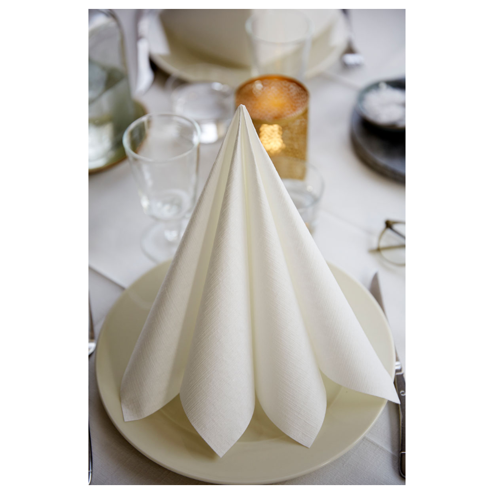 Une serviette Duni Dunilin® 48 x 48 cm blanche de Duni GmbH, soigneusement pliée, se tient debout sur une assiette blanche au milieu de verres vides, d'un chandelier rond, d'un bol de sel, de couverts et d'une nappe blanche. L'ambiance élégante souligne la texture semblable à du tissu de la serviette jetable de haute qualité.