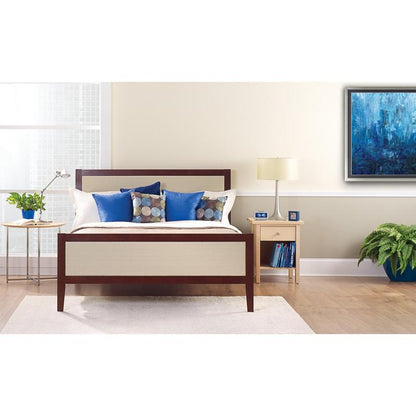 Un lit bien fait avec des oreillers blancs et bleus se trouve devant un mur clair, recouvert de ruban de masquage ScotchBlue™ "Multi-Surface" de 3M Deutschland GmbH. Sur une table de chevet, il y a une lampe, une horloge, des livres et une plante en pot à côté de la fenêtre lumineuse.