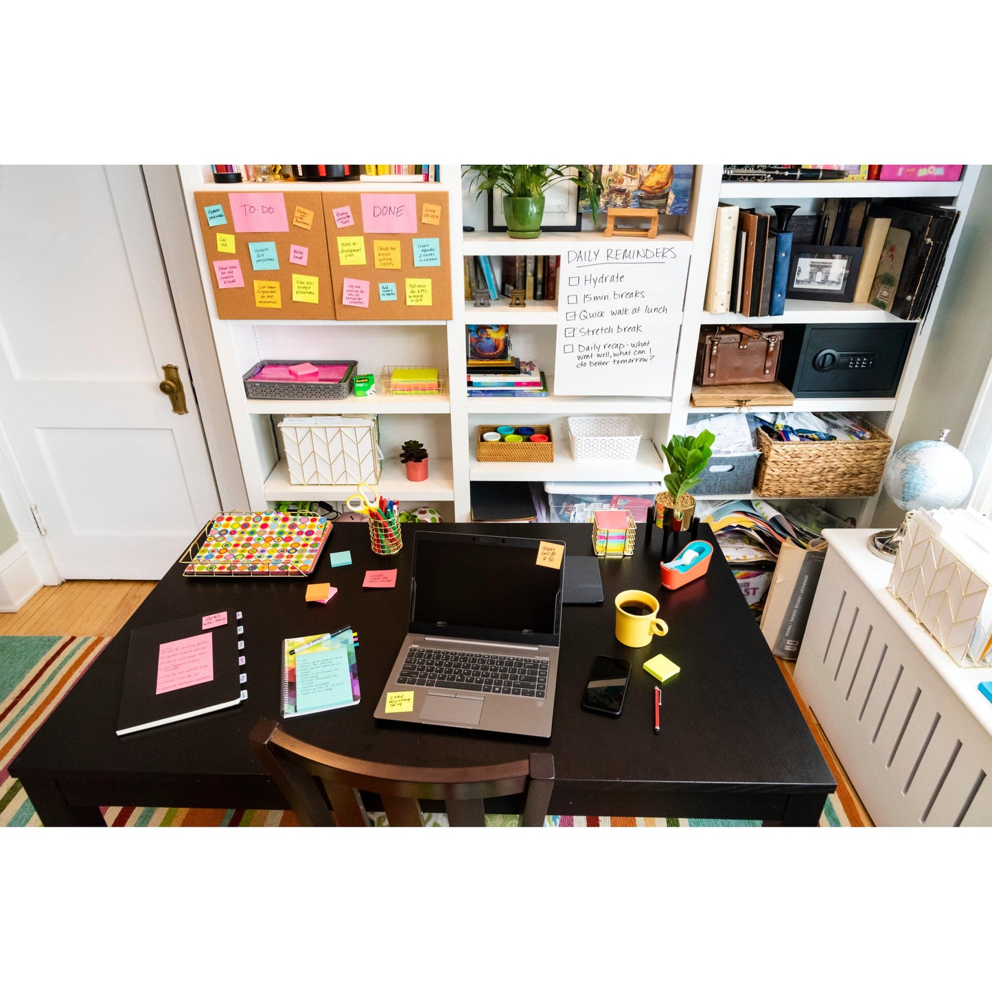 Auf dem Schreibtisch befinden sich ein Laptop, Notizbücher, ein Telefon, Stifte und eine gelbe Tasse. Post-it® Super Sticky Notes von 3M Deutschland GmbH (76 mm x 127 mm) befinden sich auf dem Schreibtisch und dem Whiteboard neben Büchern und Körben im Regal.