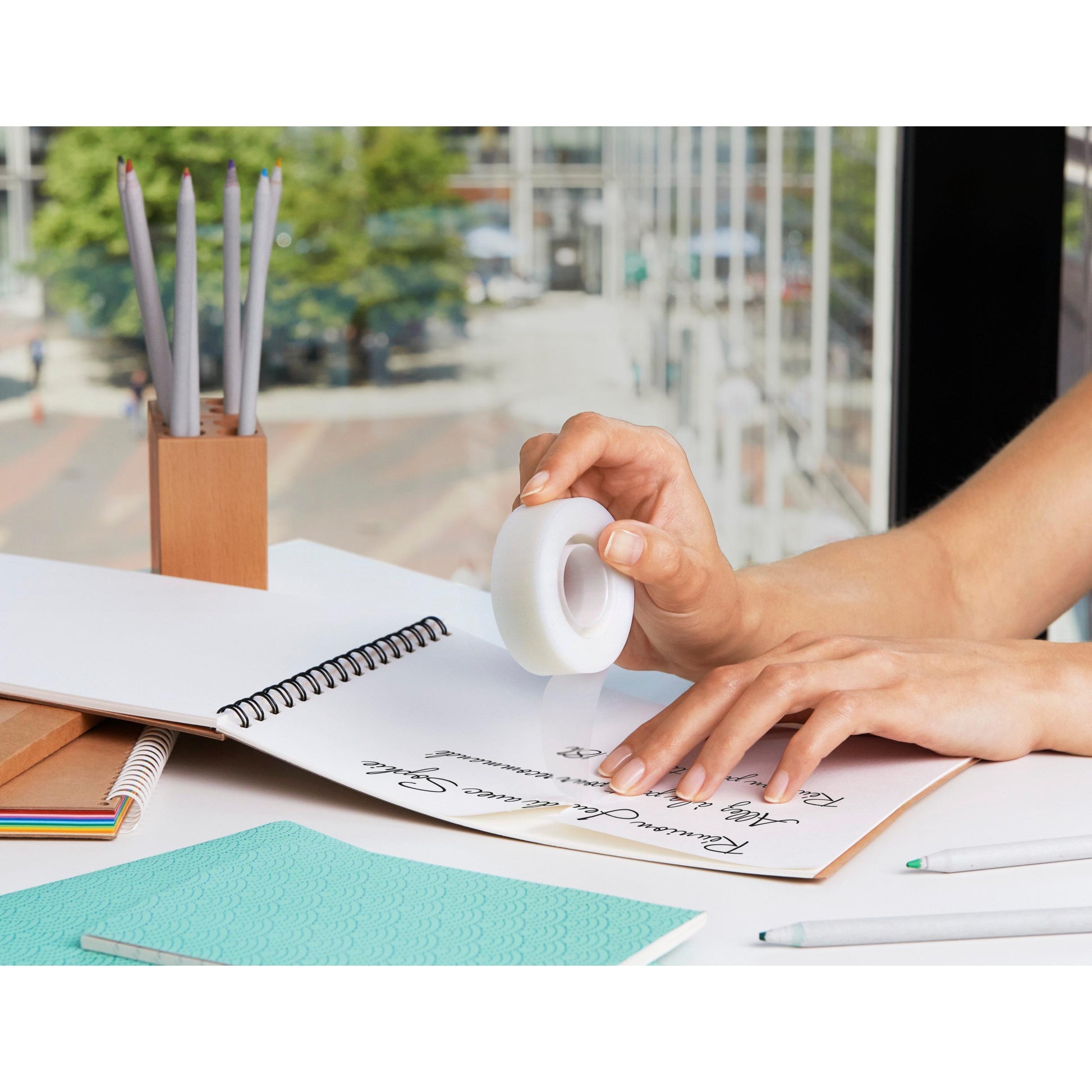 Eine Person verwendet Scotch® Magic™ Klebeband 900-1933-9 von 3M Deutschland GmbH auf handschriftlichen Notizen in einem Spiralheft. Bleistifte, ein Stifthalter und ein türkisfarbenes Notizbuch liegen auf dem Schreibtisch in der Nähe eines Fensters mit Blick auf eine Stadt und Bäume.