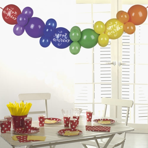 Une table blanche avec de la vaisselle de fête à pois rouges, des ustensiles jaunes et des gobelets est couronnée par la guirlande de ballons de 2 m "Happy Birthday" de PAPSTAR GmbH - une décoration de fête d'anniversaire festive qui brille au soleil à travers des volets blancs.