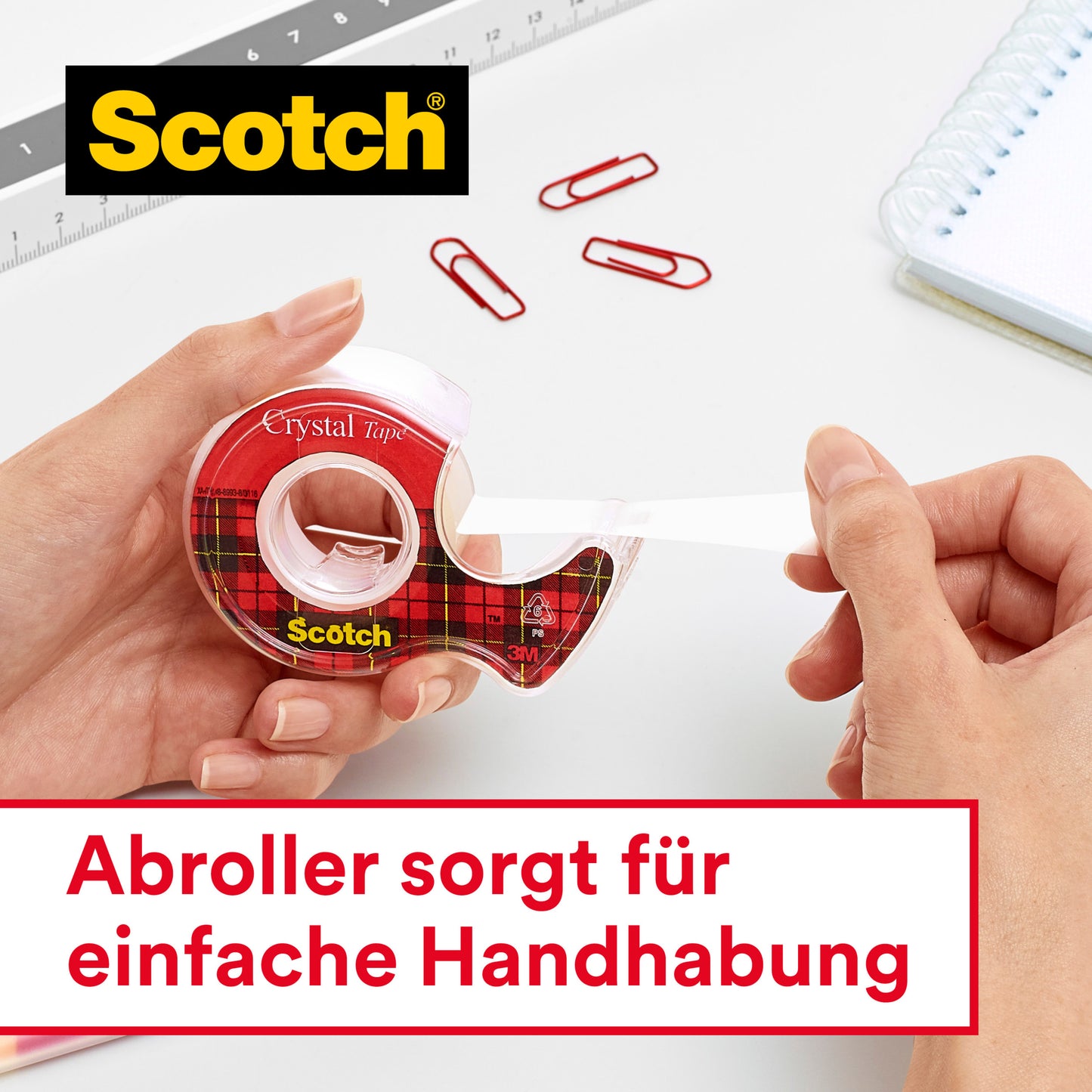 Eine Person zieht das Scotch® Crystal Klebeband (12 mm x 10 m) von 3M Deutschland GmbH aus einem Abroller. Im Hintergrund sind ein Lineal, rote Büroklammern und ein Spiralbuch. Text: "Abroller sorgt für einfache Handhabung.