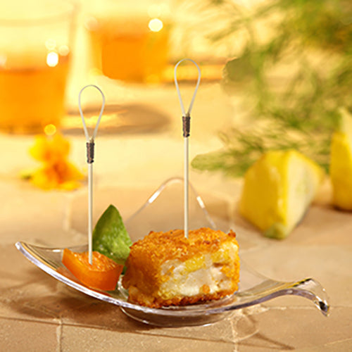 Un bol en verre présente un cube d'entrée pané et frit avec un pique à finger food "Loop" PAPSTAR (10 cm), un segment d'orange et une garniture verte. En arrière-plan, des quartiers de citron et deux boissons de la société PAPSTAR se fondent.