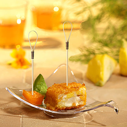 Un bol en verre présente un cube d'entrée pané et frit avec un pique à finger food "Loop" PAPSTAR (10 cm), un segment d'orange et une garniture verte. En arrière-plan, des quartiers de citron et deux boissons de la société PAPSTAR se fondent.