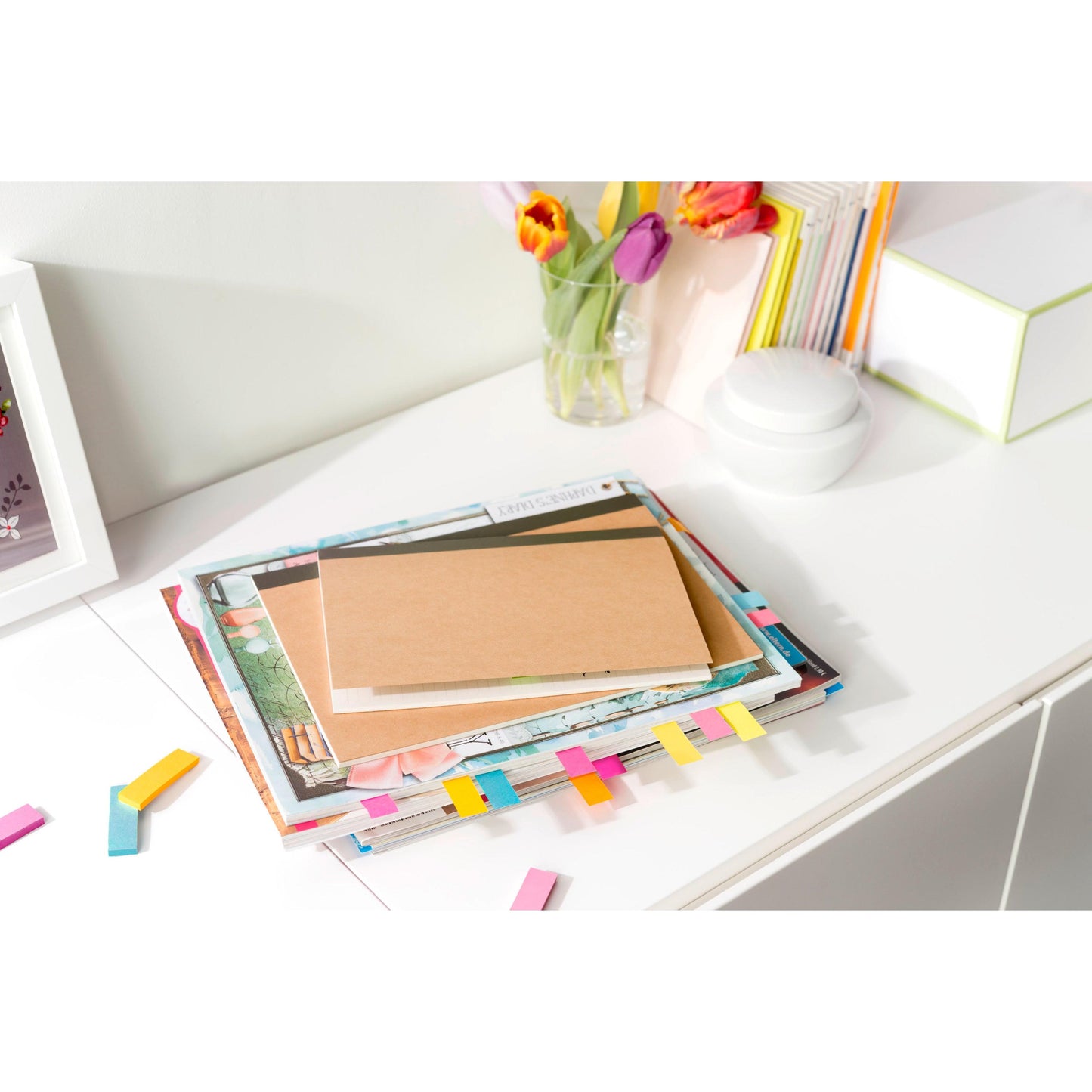 Sur un bureau blanc se trouve le marque-page Post-it® (15 mm x 50 mm, 50 feuilles/bloc, 100% PEFC) de 3M Deutschland GmbH entre des livres empilés et des carnets avec des marqueurs colorés, des notes éparpillées, un vase en verre avec des fleurs et des fournitures de bureau.