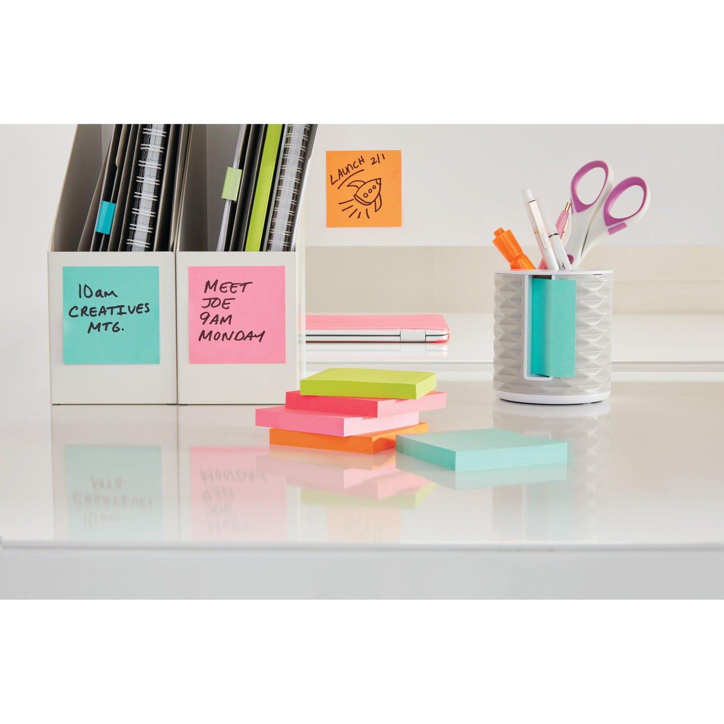 Un bureau bien rangé avec des notes colorées 3M Post-it® de la Energetic Collection (76x127 mm, 100% PEFC, SGSCH-PEFC-COC-110078) dans deux supports blancs, avec des ciseaux, des stylos et des piles rappelant des réunions et des déjeuners.