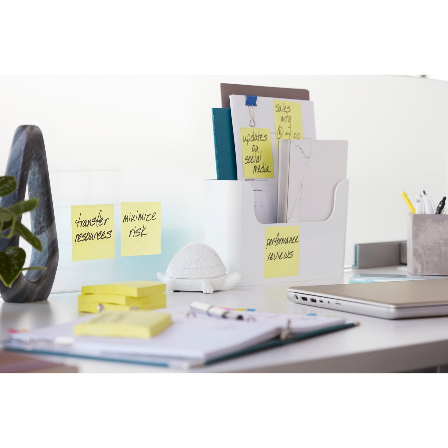Un bureau bien rangé avec des dossiers, 3M Deutschland GmbH Post-it® Super Sticky Notes (Jaune, 76x127mm, 90 feuilles/bloc, 14+2 promo-pack), un ordinateur portable, des stylos, une plante, une décoration de tortue blanche et un carnet. Les notes indiquent : "Transférer les ressources", "Minimiser les risques", "Évaluation des performances".