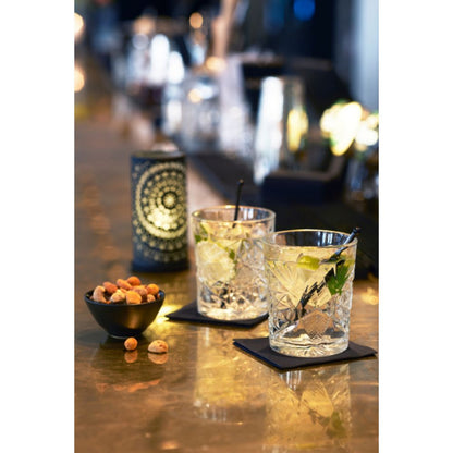 Deux verres avec de la glace, des tranches de citron vert et des pailles sont posés sur des sous-verres carrés sur un comptoir de bar. À côté, il y a un petit bol noir avec des noix mélangées. Le photophore Billy de Duni GmbH, en métal simple 140 x 75 mm au design moderne, brille doucement en arrière-plan, créant une atmosphère chaleureuse.
