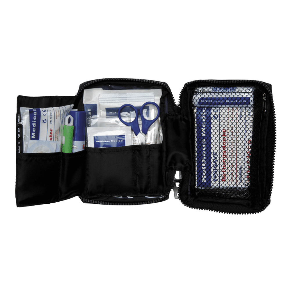 Un sac de secours Holthaus Medical TRAVEL bleu/rouge avec fermeture éclair de Holthaus Medical GmbH & Co. KG est ouvert et montre divers articles de premiers secours. Parmi les articles, on trouve des ciseaux, des pansements, des tampons alcoolisés, des compresses de gaze et un crayon vert. Une poche en filet sur le côté droit contient des pansements supplémentaires et une brochure de premiers secours pour une consultation rapide.