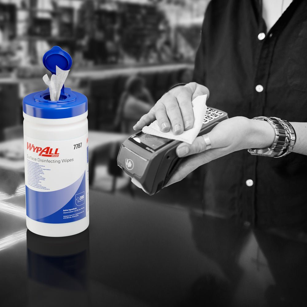 Quelqu'un utilise une lingette désinfectante WypAll® (Kimberly-Clark GmbH) d'une cuve distributrice ouverte pour désinfecter un terminal de carte. Le seau distributeur blanc (200 lingettes) est posé sur le comptoir, l'arrière-plan est flou.