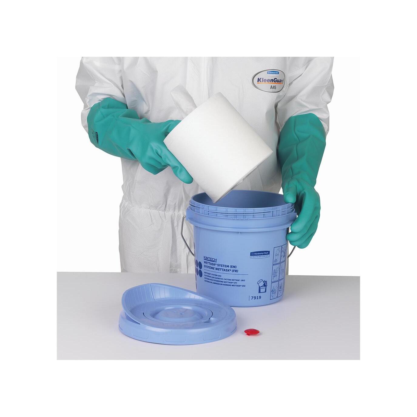 Eine Person mit grünen Handschuhen und weißem Anzug legt eine Rolle Kimtech® Wettask™-Tücher in einen blauen Spendereimer der Kimberly-Clark GmbH Wischtuch; der Deckel und die rote Kappe liegen auf dem Tisch daneben.