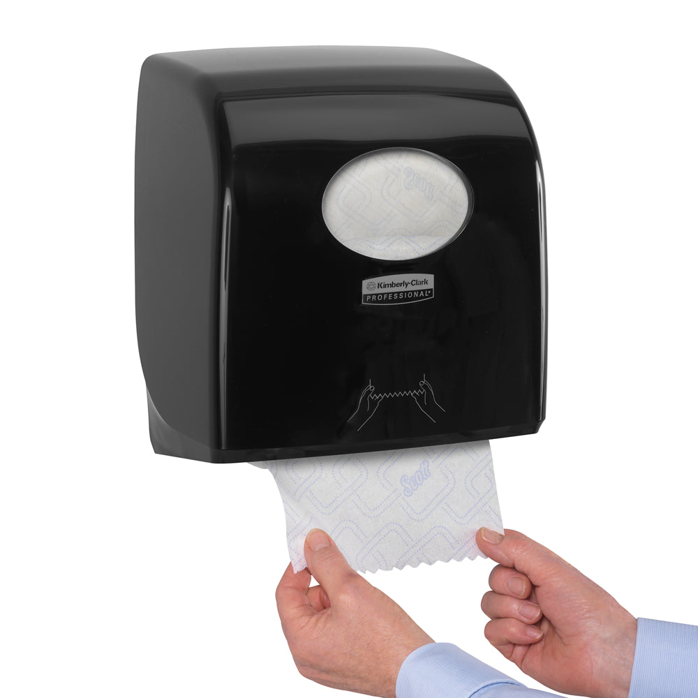 Eine Person zieht Papierhandtücher aus einem Kimberly-Clark GmbH Aquarius™ Slimroll™ Rollenhandtuchspender - Schwarz, der an der Wand montiert ist. Nur die Hände sind sichtbar und zeigen die hygienische Papierhandtuchentnahme durch das ovale Fenster.