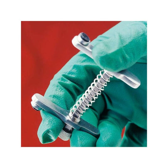 Une personne portant des gants verts tient, devant un fond rouge, un serre-mâchoires Altruan Servoprax pour chats, doté d'un manche cylindrique et d'une vis filetée, généralement utilisé par les vétérinaires pour les interventions vétérinaires.