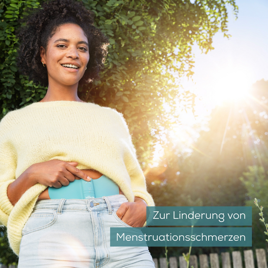 Eine Person mit lockigem Haar, einem gelben Pullover und hellen Jeans steht lächelnd im Freien mit grünem Hintergrund. Sie hält ein blaugrünes Beurer EM 55 Menstrual Relax+ Gel-Pad an ihren Bauch. Der Text auf Deutsch lautet: „Zur Linderung von Menstruationsschmerzen.