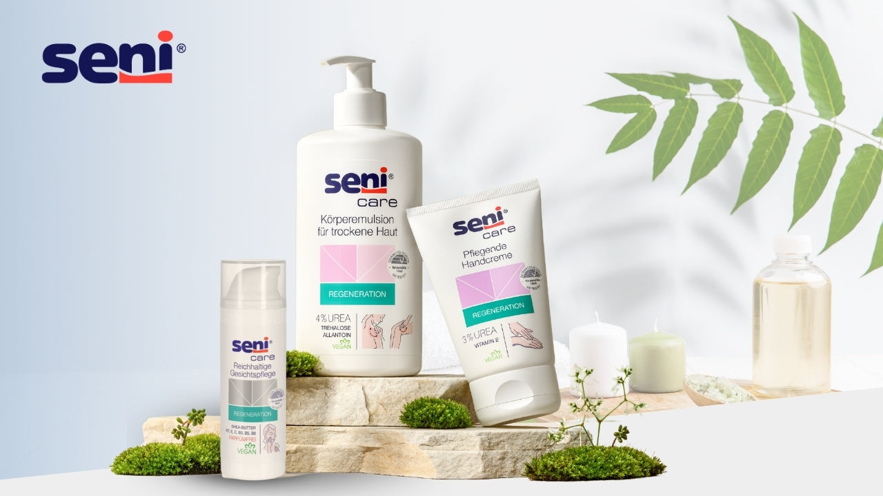 Pflegeprodukte der Marke Seni Care auf einer natürlichen Steinfläche arrangiert: eine Körperemulsion für trockene Haut, eine pflegende Handcreme und eine reichhaltige Gesichtspflege, umgeben von Moos, Pflanzen und Pflegeaccessoires vor hellem Hintergrund.