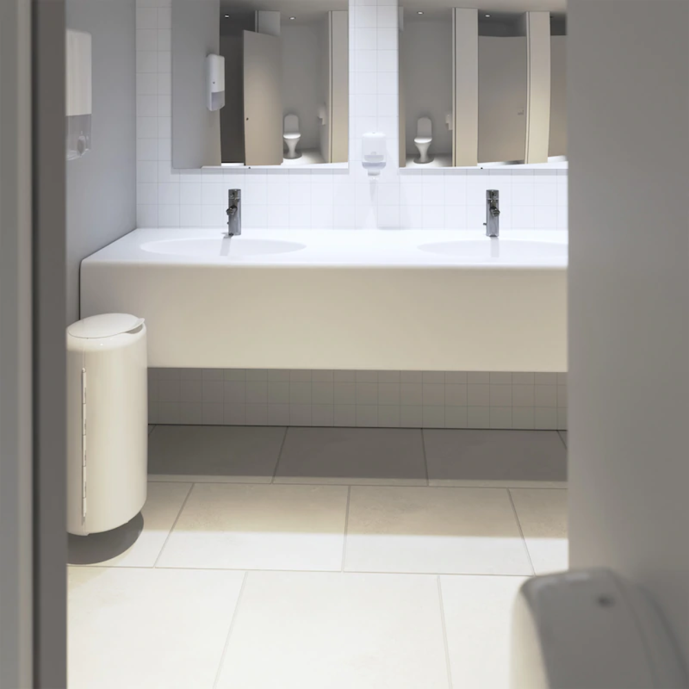La salle de bain publique moderne dispose d'un élégant plan de toilette blanc avec deux lavabos et des accessoires en chrome. À gauche se trouve une poubelle blanche, tandis que des miroirs sont suspendus au-dessus du sol carrelé. Sur le mur, des distributeurs de savon Tork Xpress® 552000 d'Essity Professional Hygiene Germany GmbH sont installés, et des serviettes pliées augmentent le confort des visiteurs.