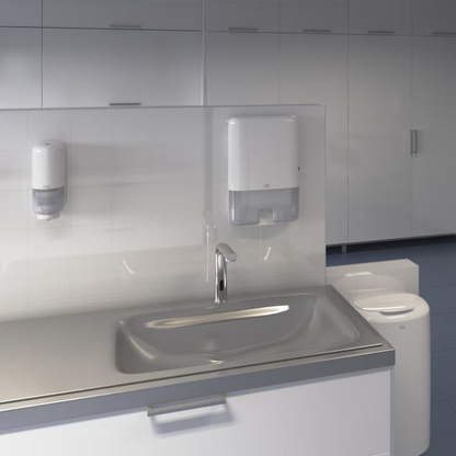 Un lavabo moderne est montré dans des toilettes publiques avec un robinet automatique. Au-dessus du lavabo, un distributeur Tork Xpress® 552000 pour serviettes pliées en H2 d'Essity Professional Hygiene Germany GmbH et un distributeur de savon sont montés sur un mur carrelé. En arrière-plan, une rangée d'armoires blanches est visible.