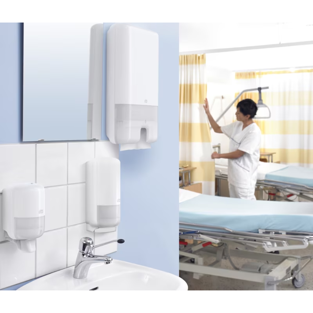 Dans une chambre d'hôpital, un médecin en uniforme blanc se tient à côté d'un lit. Au premier plan se trouve un lavabo équipé d'un distributeur Tork Xpress® 552000 pour serviettes pliées en H2 de TORK - Essity Professional Hygiene Germany GmbH. La chambre est équipée de dispositifs médicaux et de rideaux rayés jaunes.