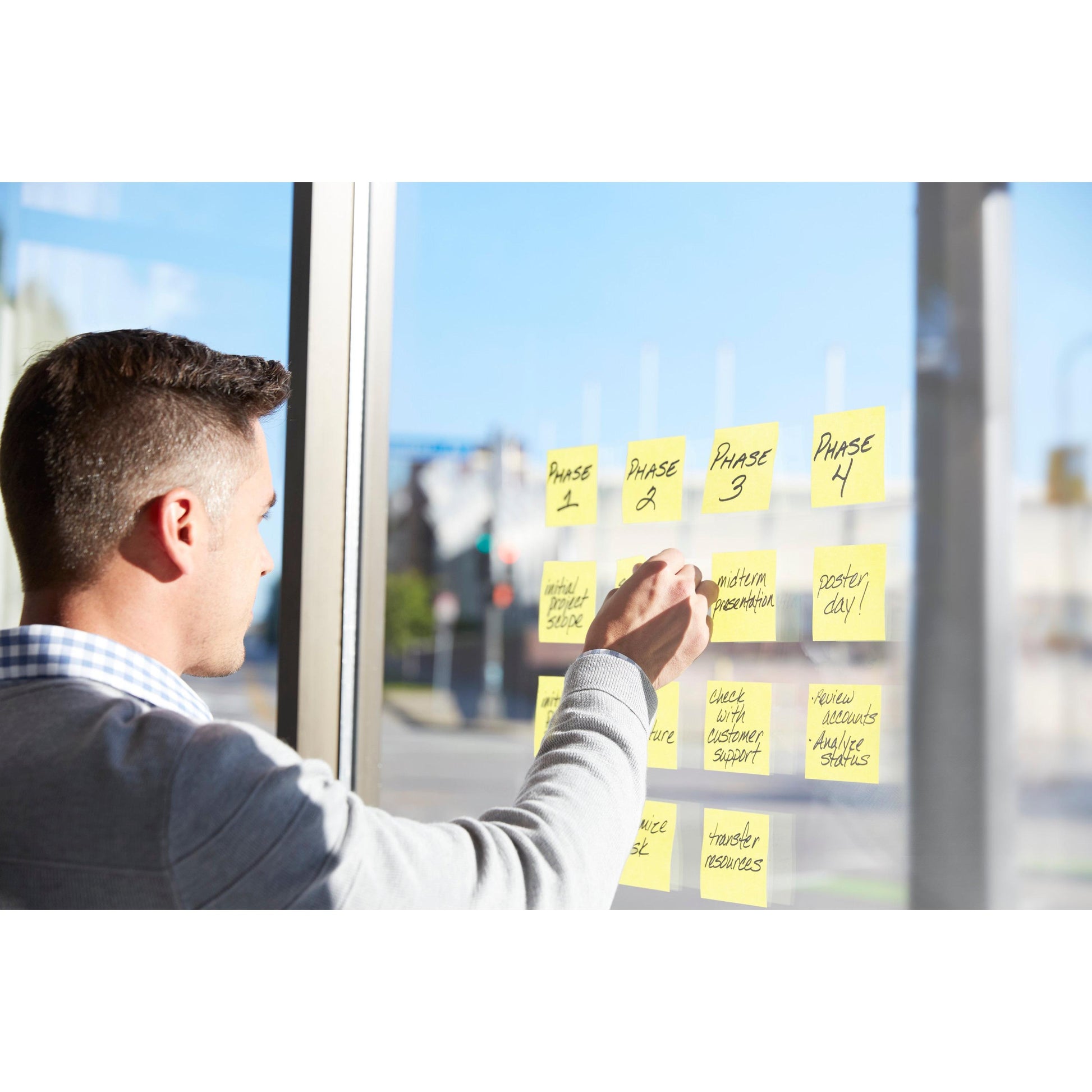 Eine Person benutzt Post-it® Super Sticky Notes, Gelb, 76x76 mm (3M Deutschland GmbH), in Innenräumen, um Projektphasen an einem Glasfenster mit Blick auf eine städtische Straße zu organisieren. Aktionspackung: 14 Blöcke + 2 gratis, 100% PEFC-zertifiziert.