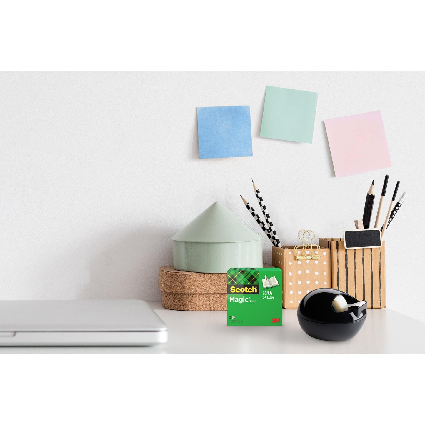 Ein Schreibtisch mit einem geschlossenen Laptop, 3M Scotch™ Magic Tape, ein schwarzer Scotch™ Karim Rashid Tischabroller von 3M Deutschland GmbH, Becher mit Stiften und Bleistiften, gestapelte runde Schachteln und drei pastellfarbene Haftnotizen an einer weißen Wand.