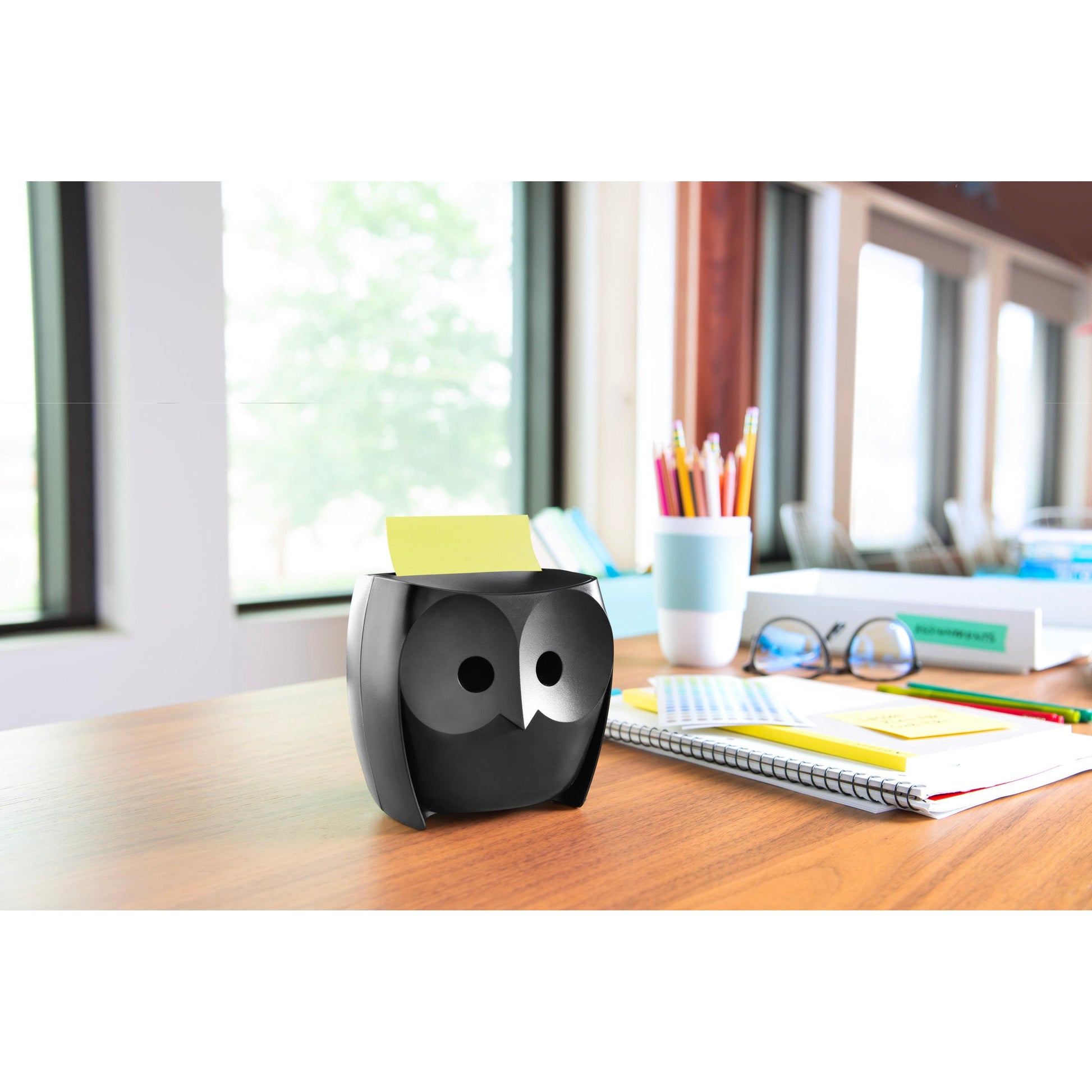Der 3M Deutschland GmbH Post-it® Z-Notes Dispenser, Eule, Schwarz, mit Post-it® Super Sticky Z-Notes (45 Blatt/Block, 2 Blöcke/Packung), steht auf einem Holztisch neben Notizbüchern und Stiften in einem hell erleuchteten Raum.