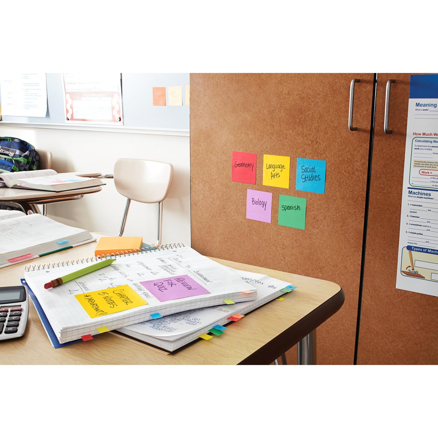 Représentée est une salle de classe avec des cahiers, une calculatrice et des papiers. Sur l'armoire se trouvent des Post-it® Super Sticky Notes (76x76 mm, 90 feuilles/bloc, 100% PEFC) de 3M Deutschland GmbH, étiquetés pour différentes matières; à l'arrière-plan, on voit une chaise vide et des affiches.