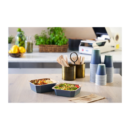 Auf dem Tisch stehen zwei Take-away-Schüsseln, flankiert von gestapelten Duni Urban Kaffeebechern PAP/PLA der Duni GmbH, Holzbesteck in einem Halter und Servietten. Zitronen, Gläser und Kräuter im Hintergrund tragen zum nachhaltigen Ambiente dieses Essenszubereitungsbereichs bei.